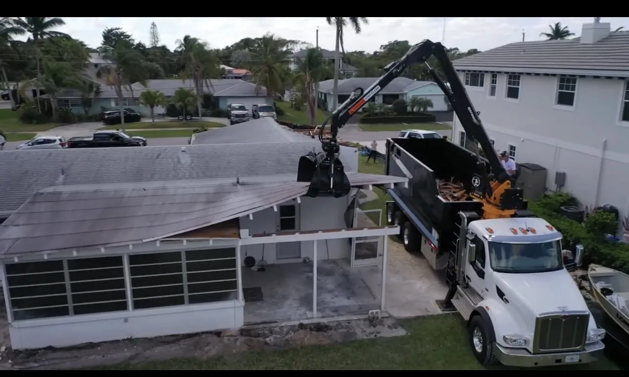 Grapple truck with its hydraulic loader extended beside a partially dismantled residential structure